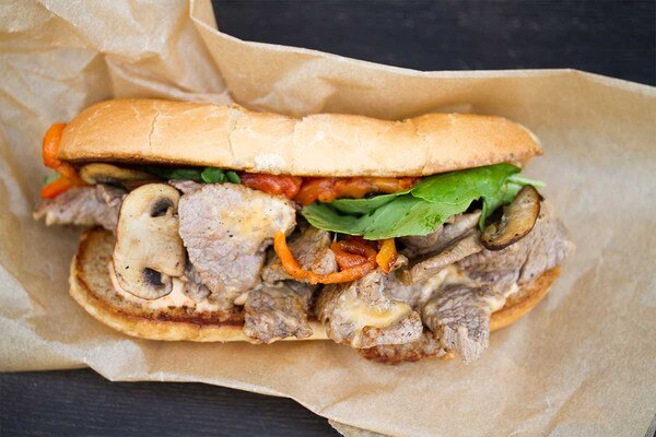 Philly cheese steak sandwich with grain-fed veal stew meat, mushrooms, lettuce, red bell pepper on brown butcher paper, black background