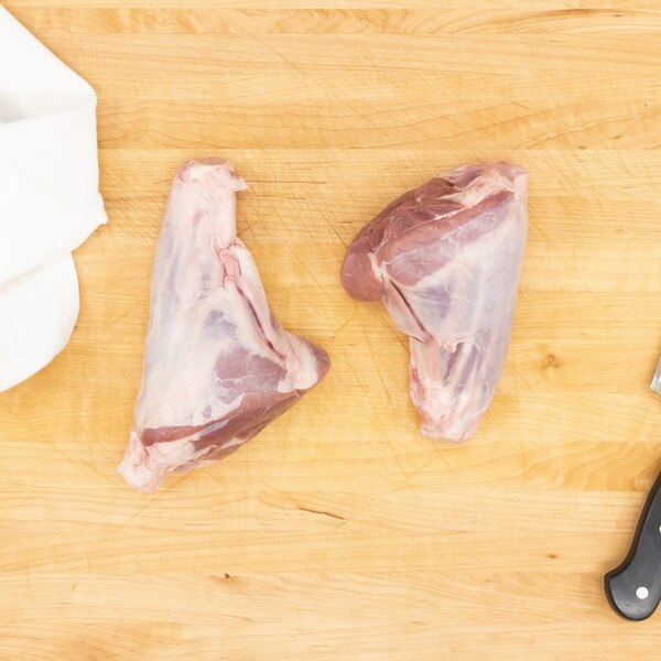A pair of raw, Welsh lamb hindshanks on a wooden board with a crumpled white towel to the left and a black knife handle to the right.