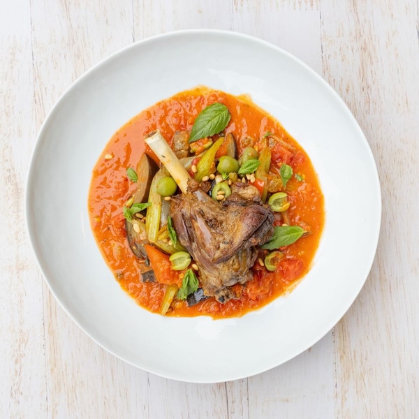 On a whitewashed wood background, a single, braised Welsh lamb hindshank sits in a round white bowl surrounded by tomato sauce with green olives, celery, pine nuts, and basil leaves.
