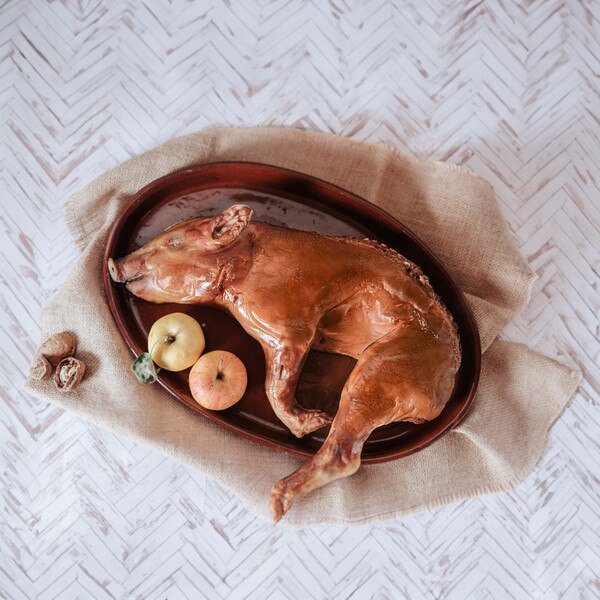 A whole, roasted suckling pig laid on its side alongside two apples and some walnuts on a glazed terra cotta oval platter cushioned by muslin against a herringbone weathered-wood background.