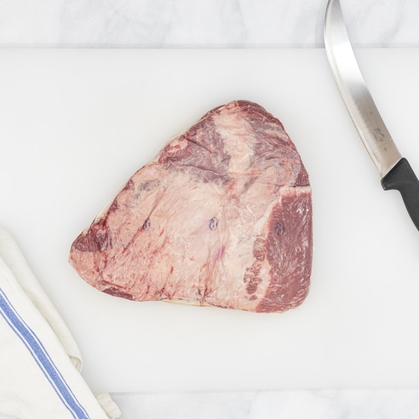 A raw Ocean Beef chuck rib on a white cutting board flanked by a blue-striped, white towel on the left and a chef’s knife on the right.