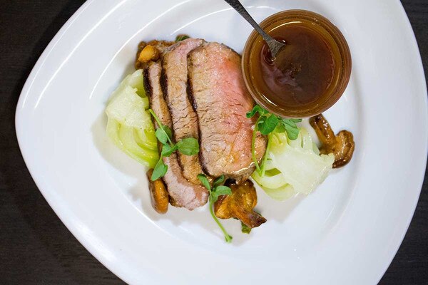 Cooked, sliced grain-fed veal striploin, bowl of fish sauce caramel with spoon, bok choy, mushrooms, white triangular plate