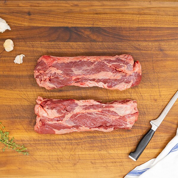 Two grass-fed beef hanging tenders on a wooden board with a knife, blue-striped kitchen towel, herb sprigs and garlic cloves.