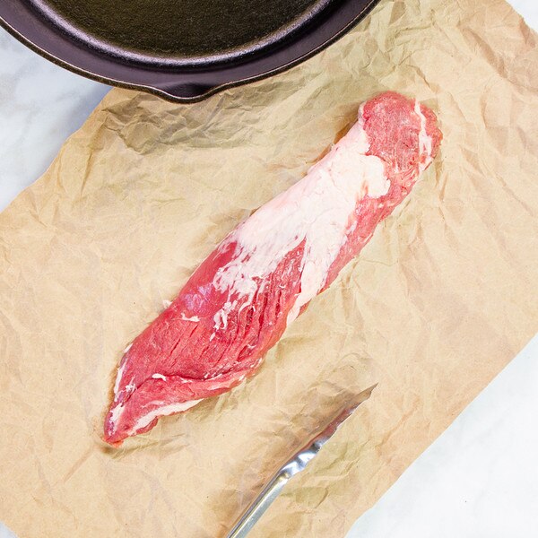 Raw, whole Spanish Iberico pork tenderloin on brown butcher paper with black skillet & silver tongs