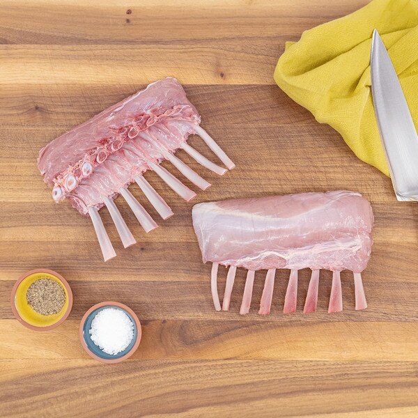 Two raw petite veal racks on a wood cutting board with bowls of salt and pepper, and a chef’s knife on a yellow towel