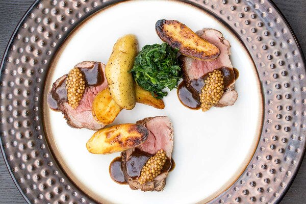 3 slices of cooked grain-fed veal butt tender, fingerling potatoes, black garlic sauce, mustard seeds, mustard greens, white plate with dotted bronze rim