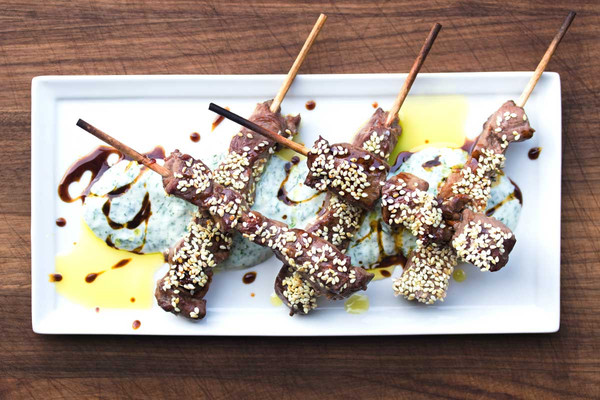5 cooked venison skewers, crisscrossed, sesame-crusted, white tzatziki sauce, pomegranate molasses drizzle, white rectangular plate, brown-stained wood background