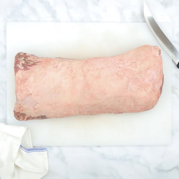 A whole, raw, boneless striploin made of barley-fed wagyu beef sits on a white cutting board against a marble background with a blue-striped, white towel to the left and a chef’s knife to the righ