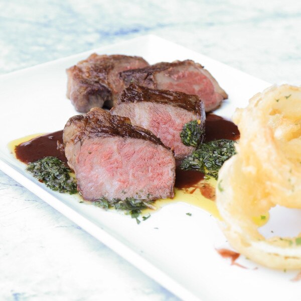 Slices of cooked barley-fed wagyu beef striploin on a rectangular white plate with a brown sauce, an herb sauce, and a side of onion rings.