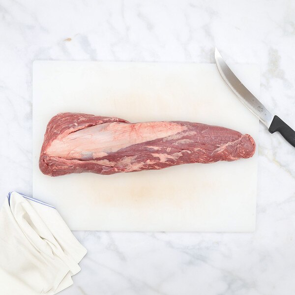 Whole, raw, barley-fed wagyu beef tenderloin on a white cutting board with a blue-striped, white towel at the bottom left, and a knife at the top right.