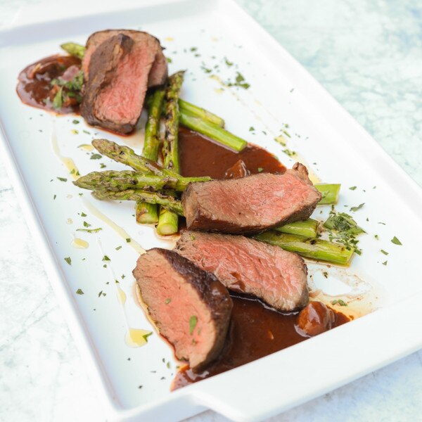 Cooked slices of barley-fed wagyu beef tenderloin with brown sauce, asparagus and minced green herbs on a rectangular white plate.