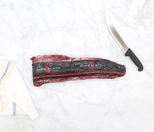 Vacuum-packed whole, barley-fed wagyu beef tenderloin with a Pure Black label on a marble background with a blue-striped, white towel at the bottom left, and a knife at the top right.