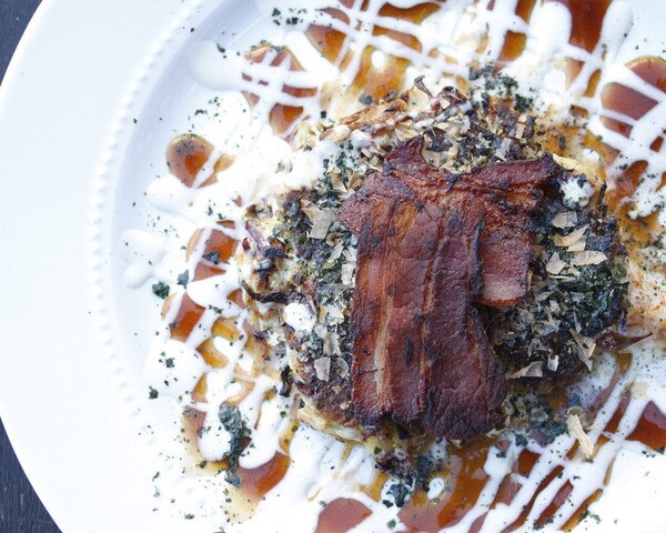 Cooked slices of bacon over a savory pancake with a crisscross pattern of white & brown sauces on a white plate