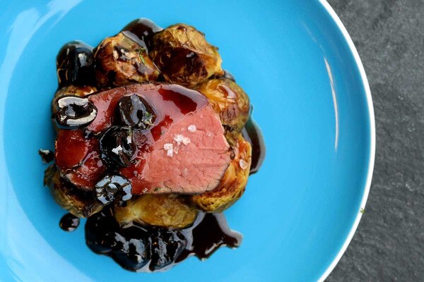 Cooked slice of bison tenderloin, cherry sauce, roasted brussels sprouts, blue plate on gray concrete background