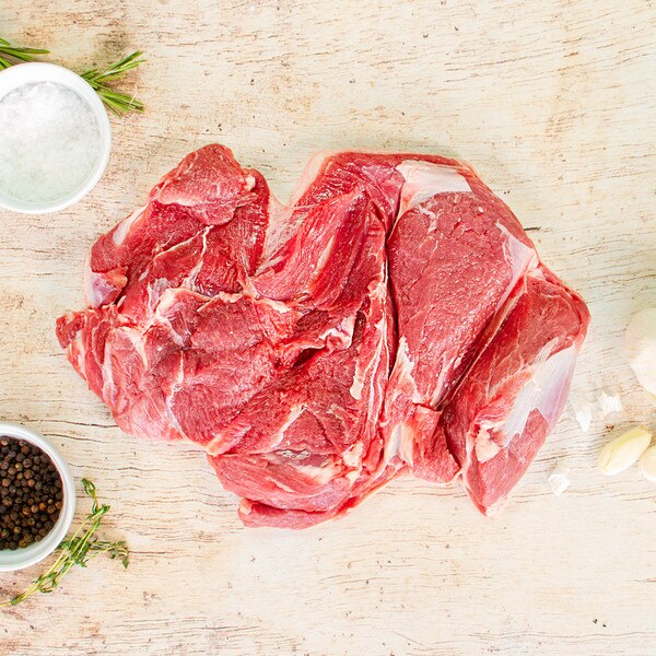 Raw boneless grass-fed lamb shoulder on whitewashed wood with bowls of salt & pepper, herb springs & garlic cloves