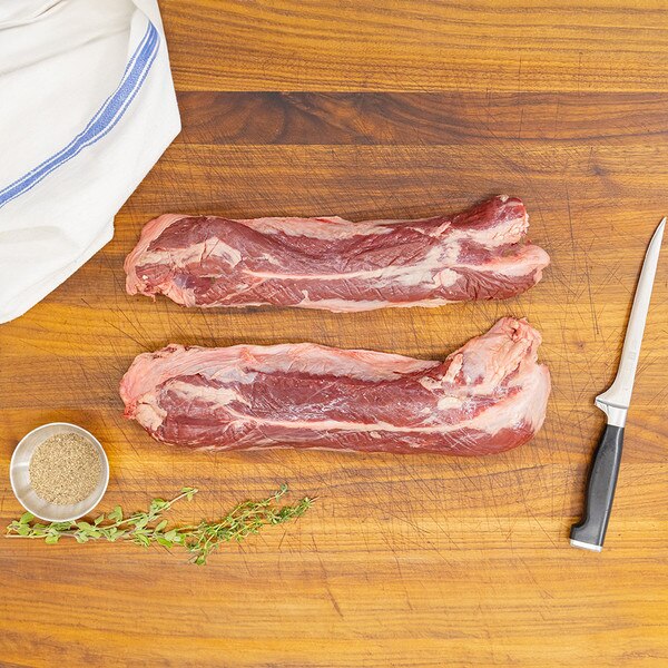 Two raw, grain-fortified beef hanger steaks on a wooden board with a knife, herb sprigs, bowl of seasoning and blue-striped kitchen towel.