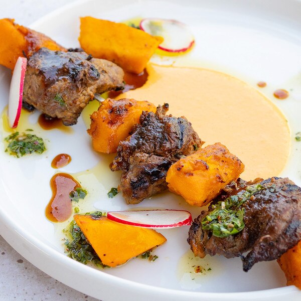 Alternating pieces of red kuri squash and oyster cut steak tips cooked on a skewer surrounded by radish rounds, dots of green herb sauce and brown demi-glace, and a golden pool of red pepper aioli.