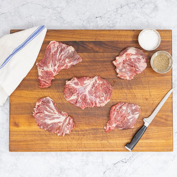 Five raw oyster cut steaks arranged on a wooden cutting board surrounded by a carving knife, a blue-striped, white kitchen towel, and silver bowls of salt and pepper.