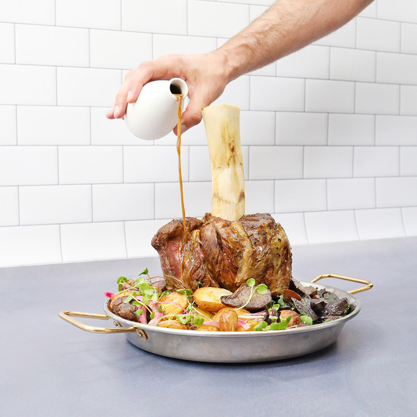 Against a white-tiled background, a hand pours gravy over Thor’s Hammer, a bone-in shank of grain-fortified beef that sits amid roasted potatoes, red onion, and microgreens in a silver pan.