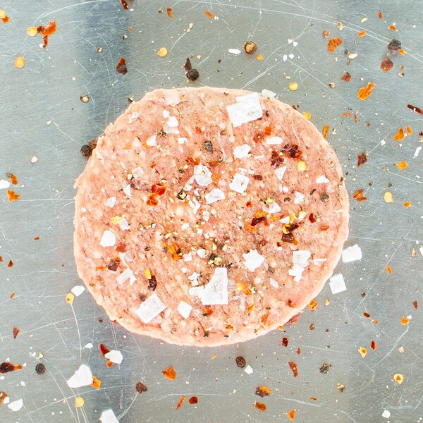 Raw grass-fed lamb burger patty on metal sheet pan sprinkled with salt & red pepper