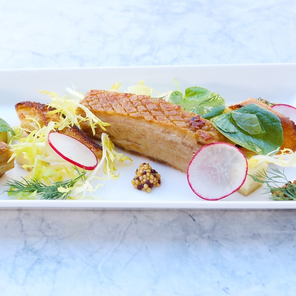 A slice of cooked Iberico pork belly filet on a rectangular white plate against a marble background, flanked by radish rounds, herb sprigs, leafy greens, and pickled mustard seeds.