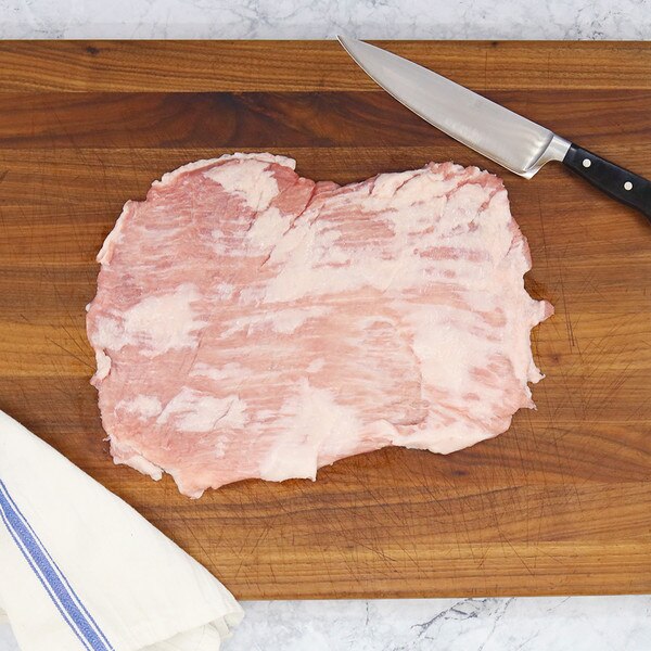 Top-down view of one whole raw Iberico pork belly filet on a wooden board against a marble background with a blue-striped, white towel in the bottom left, and a chef’s knife, top right.