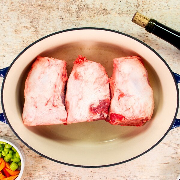 3 raw, Kurobuta pork hindshanks in an oval pot, bowl of carrots & celery, corked wine bottle on whitewashed wood