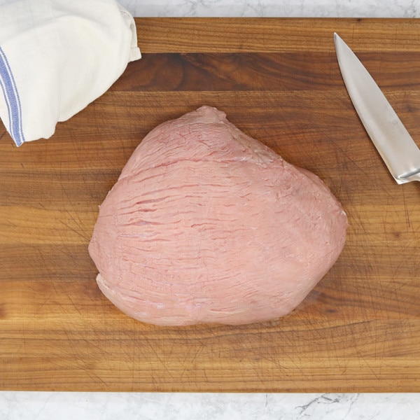 A raw, milk-fed French veal cap off top round on a wooden board with a blue-striped white towel to the left and a chef’s knife on the right.