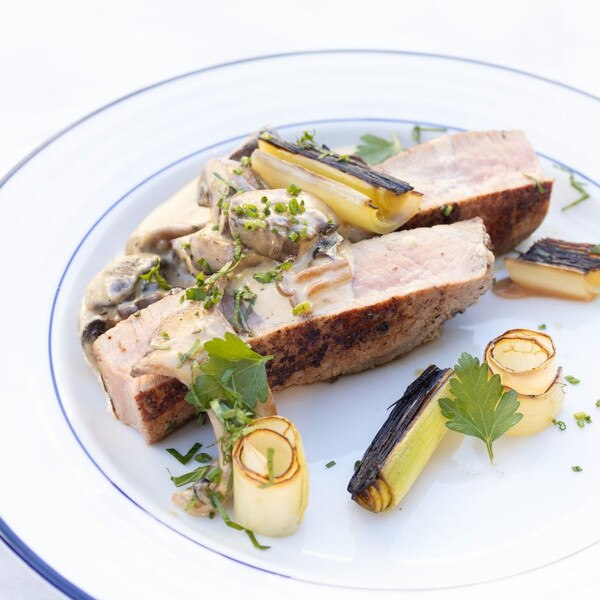 Pan-roasted milk-fed veal top round steak cut in two pieces, with creamed mushrooms, charred leeks & parsley sprigs on a blue-banded white plate.