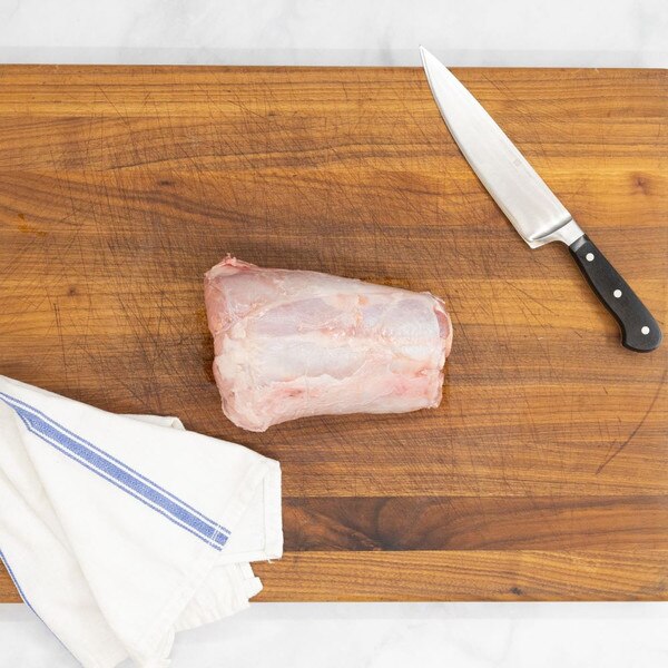 A raw, milk-fed veal center-cut hindshank on a wooden cutting board with a blue-striped, white towel on the lower left and a knife on the upper right.