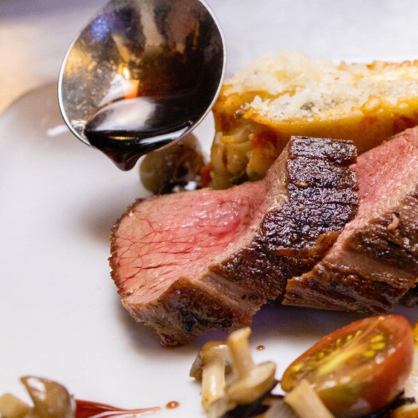 A silver ladle of brown gravy poised over slices of cooked venison leg on a white plate, mushrooms and a cherry tomato in the foreground, mushroom lasagna in the background