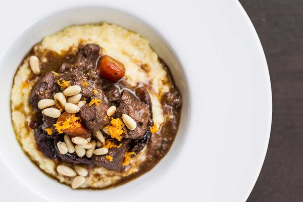 A wide-rimmed white bowl holds venison stew with white beans and carrots, served over creamy polenta.