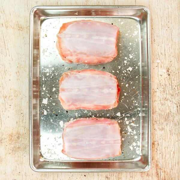 3 bone-in rabbit saddles sprinkled with salt & pepper in silver sheet pan on whitewashed wood background